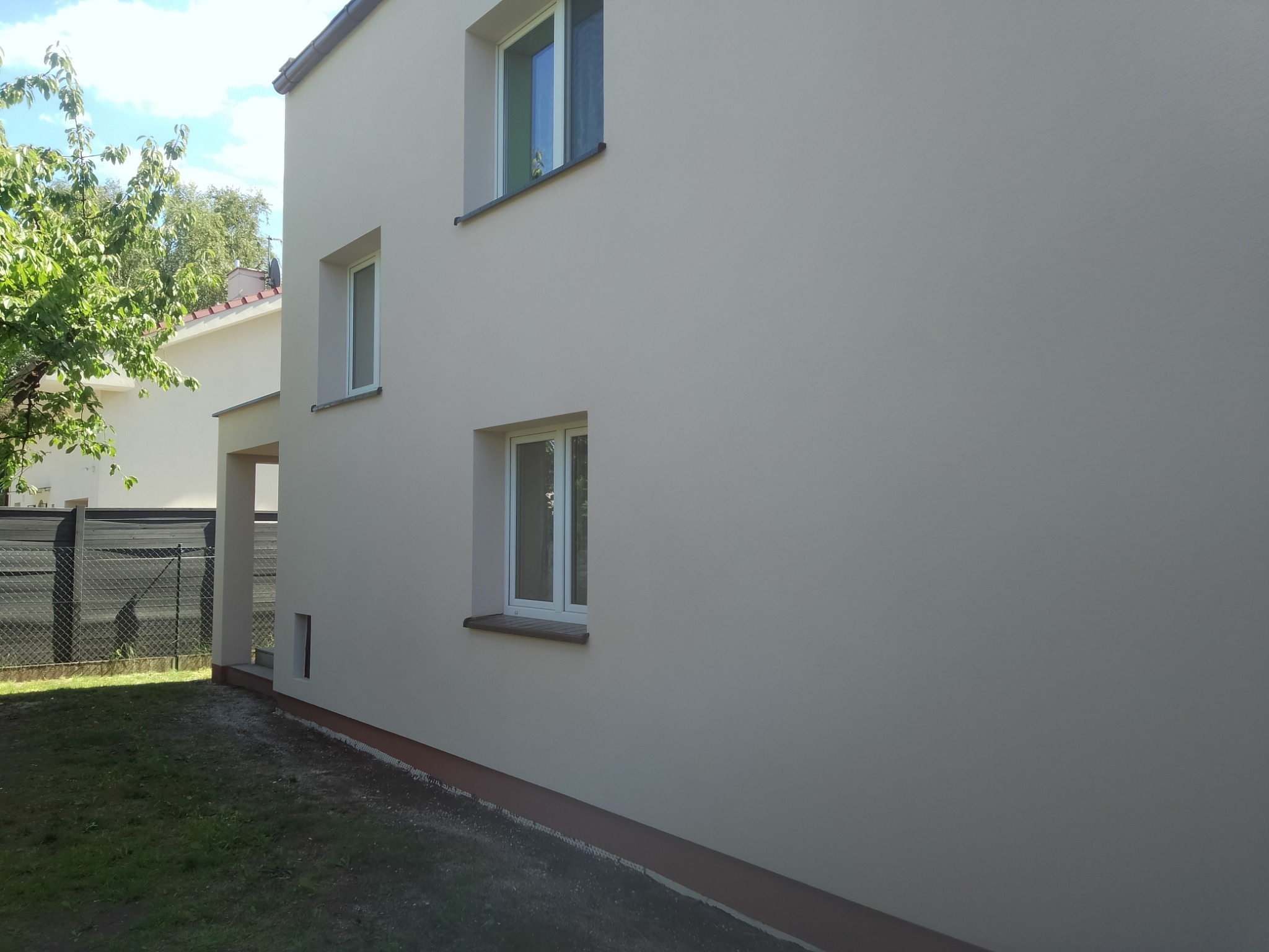 Świeżo pomalowana elewacja domu jednorodzinnego w jasnym odcieniu beżu, z trzema oknami o białych ramach i brązowym cokołem przy ziemi, widok z boku na tle zieleni.