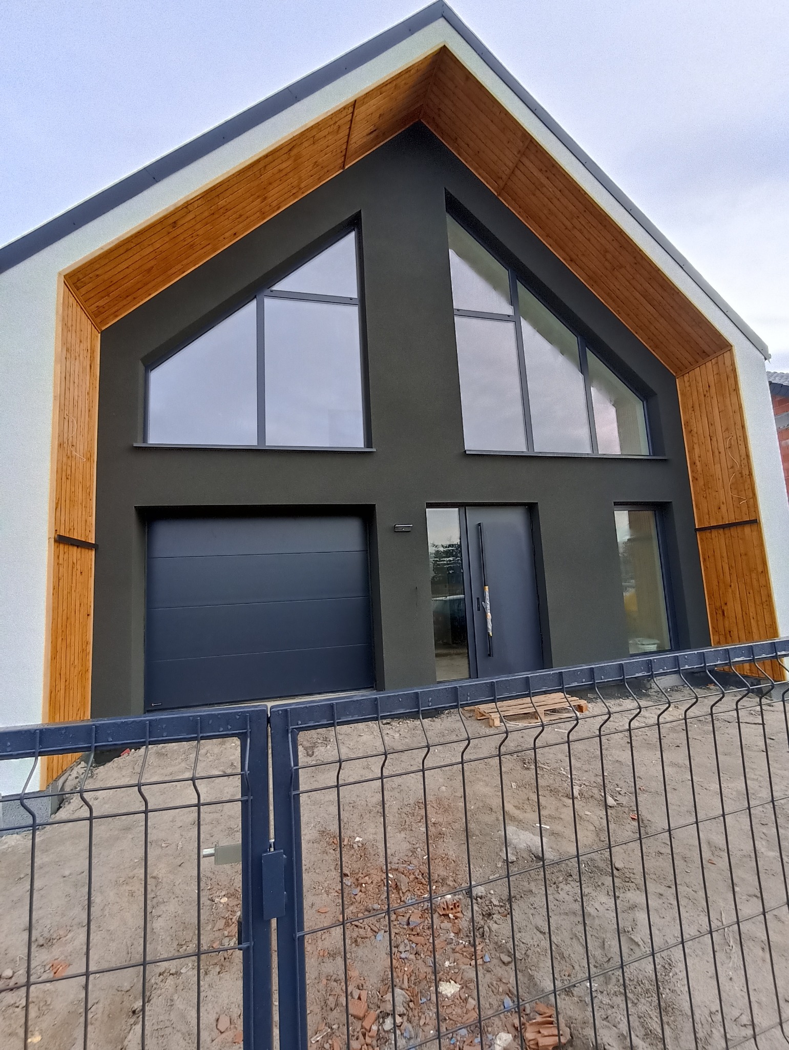 Nowoczesny dom jednorodzinny z grafitową elewacją, drewnianymi akcentami i dużymi oknami trójkątnymi, widziany zza metalowego ogrodzenia.