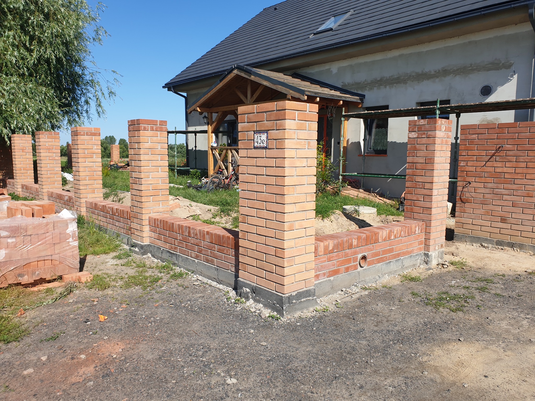 Ceglany murek z filarami w trakcie budowy przed domem z szarą elewacją i czarnym dachem. Widoczne rusztowania i materiały budowlane.