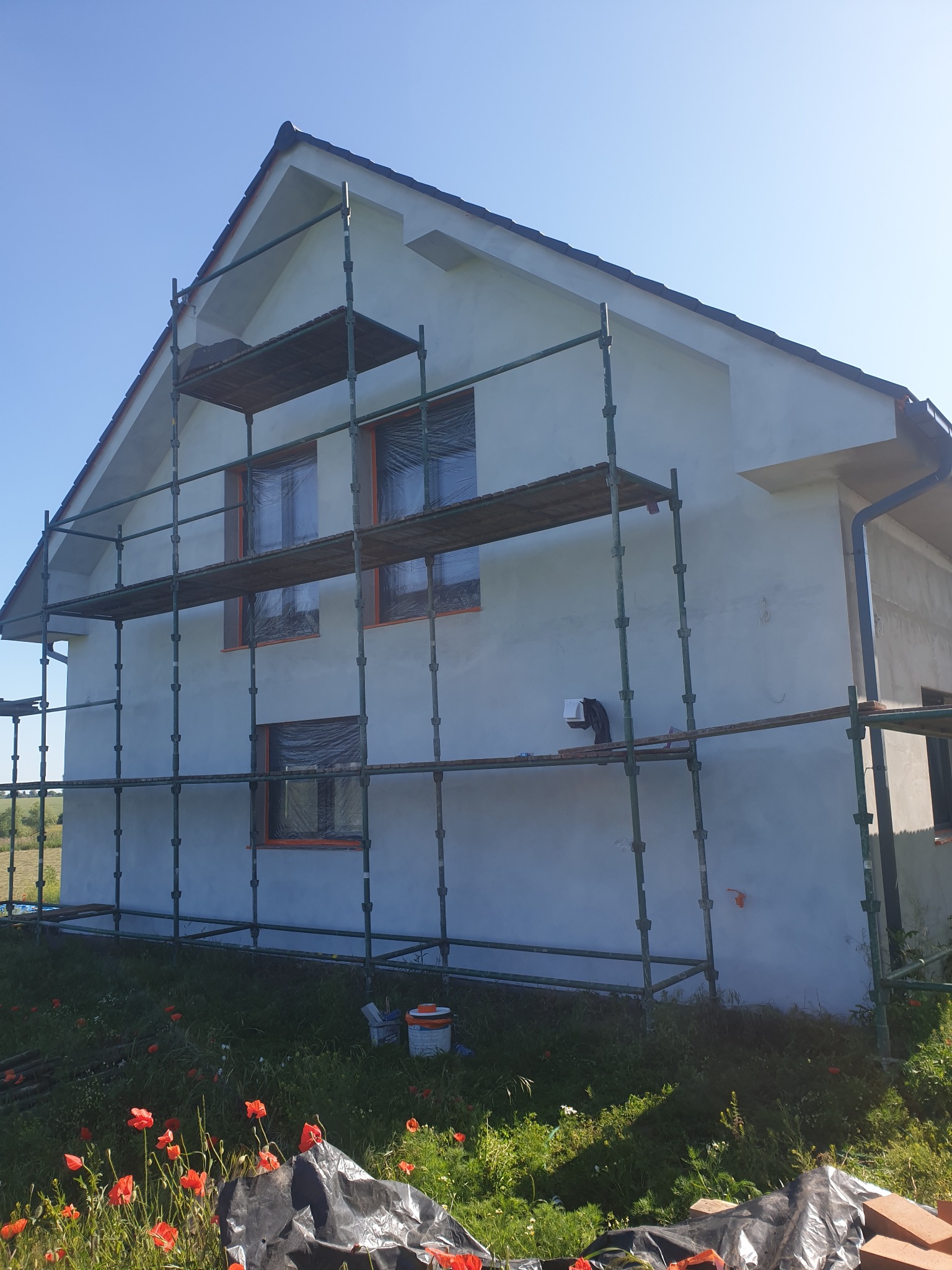 Dom w trakcie ocieplania, otoczony rusztowaniem. Okna zabezpieczone folią, widoczne farby i narzędzia. W tle łąka z makami, słoneczny dzień.