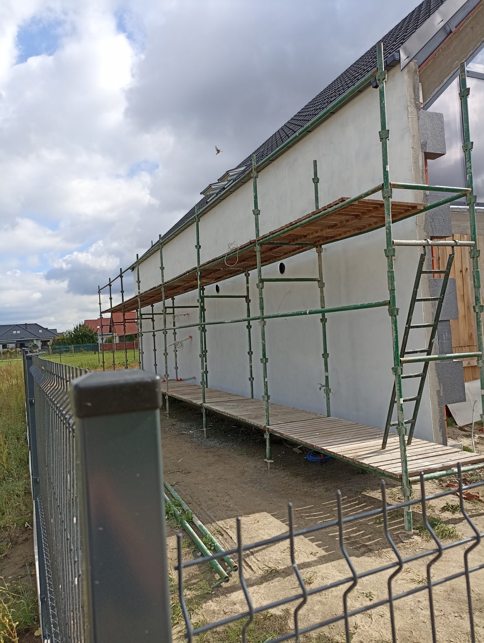 Świeżo otynkowana ściana domu jednorodzinnego z zielonym rusztowaniem i drewnianymi pomostami, widok zza metalowego ogrodzenia. W tle błękitne niebo z chmurami.
