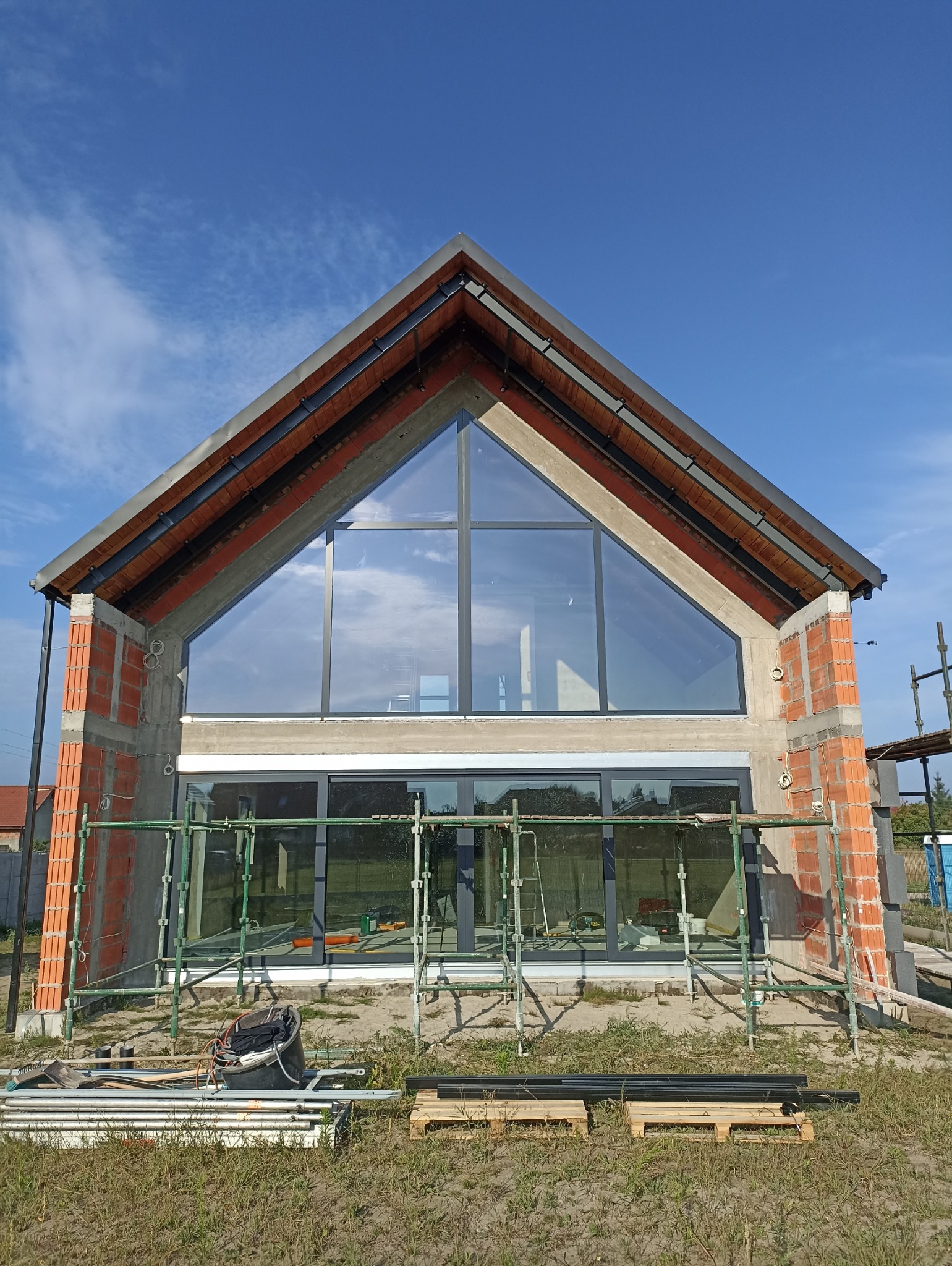 Dom w trakcie budowy z dużymi oknami i rusztowaniami. Widoczne cegły i beton. Błękitne niebo w tle. Etap surowy zamknięty, przed ociepleniem.