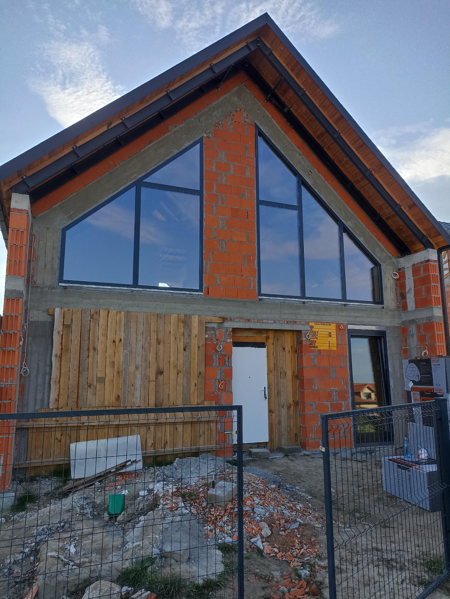 Surowy stan domu jednorodzinnego z czerwonej cegły z zamontowanymi oknami o nietypowym kształcie, zabezpieczony drewnianą konstrukcją, widok zza metalowego ogrodzenia.