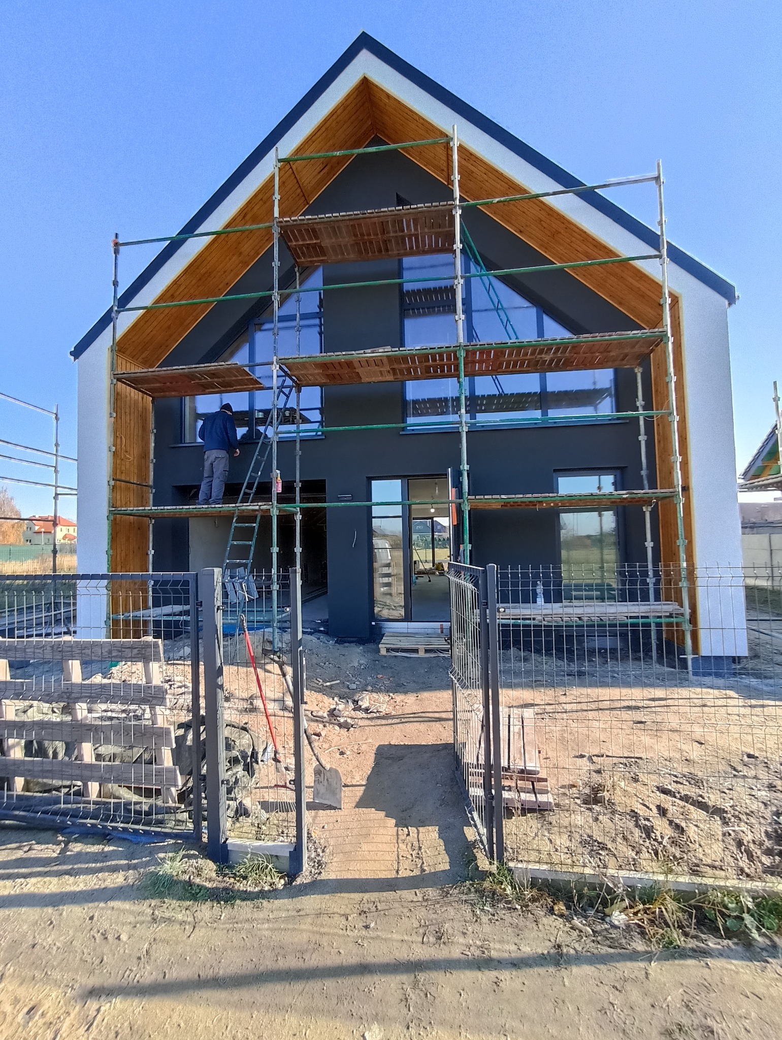 Nowoczesny dom w trakcie malowania elewacji na szaro, z drewnianymi elementami wykończeniowymi. Widoczne rusztowania i pracownik na drabinie. Błękitne niebo w tle.