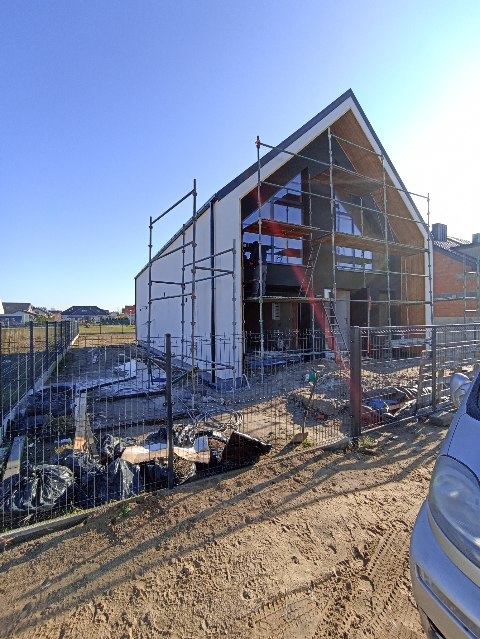 Dom jednorodzinny w trakcie ocieplania, widoczne rusztowania i częściowo otynkowana elewacja. W tle błękitne niebo i inne domy.