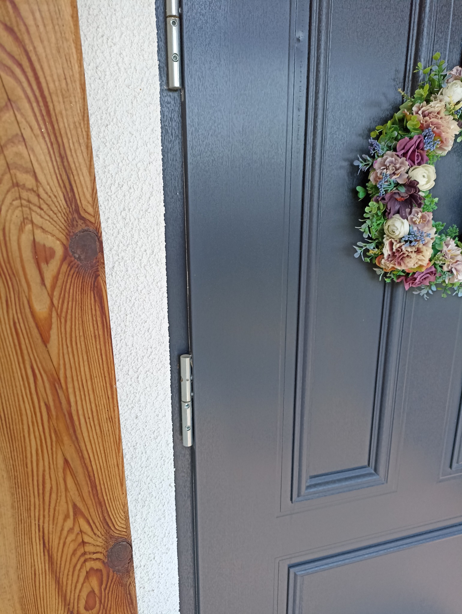 Ciemnoszare drzwi wejściowe z dekoracyjnym wieńcem kwiatowym, widoczne zawiasy i fragment drewnianej boazerii na tle białego, chropowatego tynku.