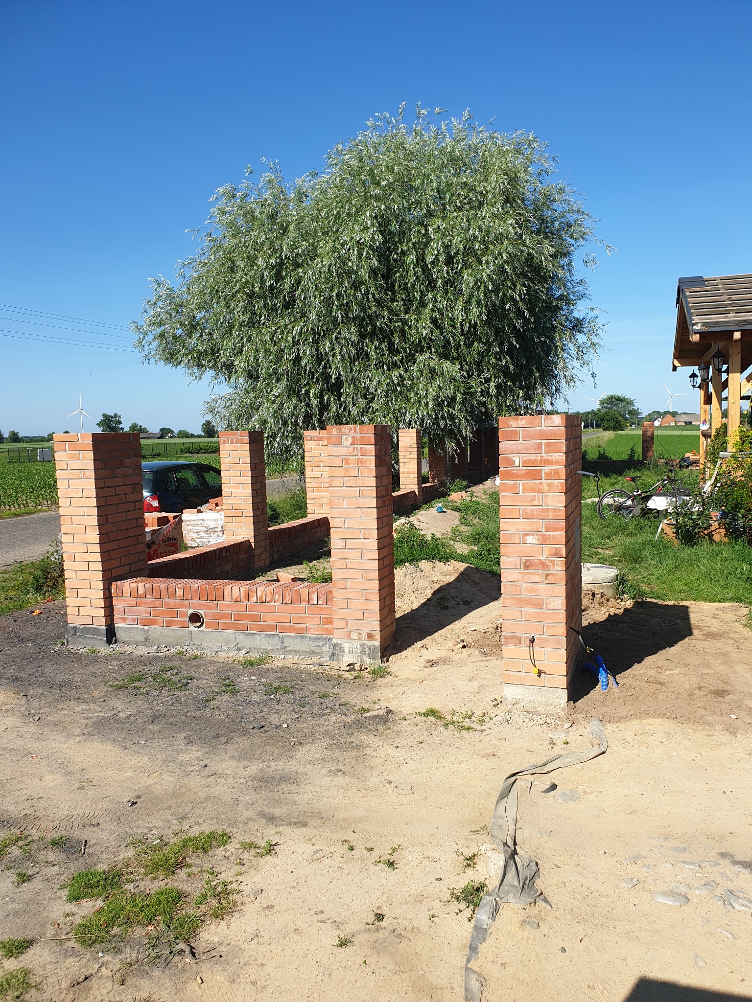Murowana konstrukcja z czerwonej cegły w trakcie budowy, widok na zielone pole i błękitne niebo. Częściowo widoczny samochód i drzewo. Prace murarskie w toku.