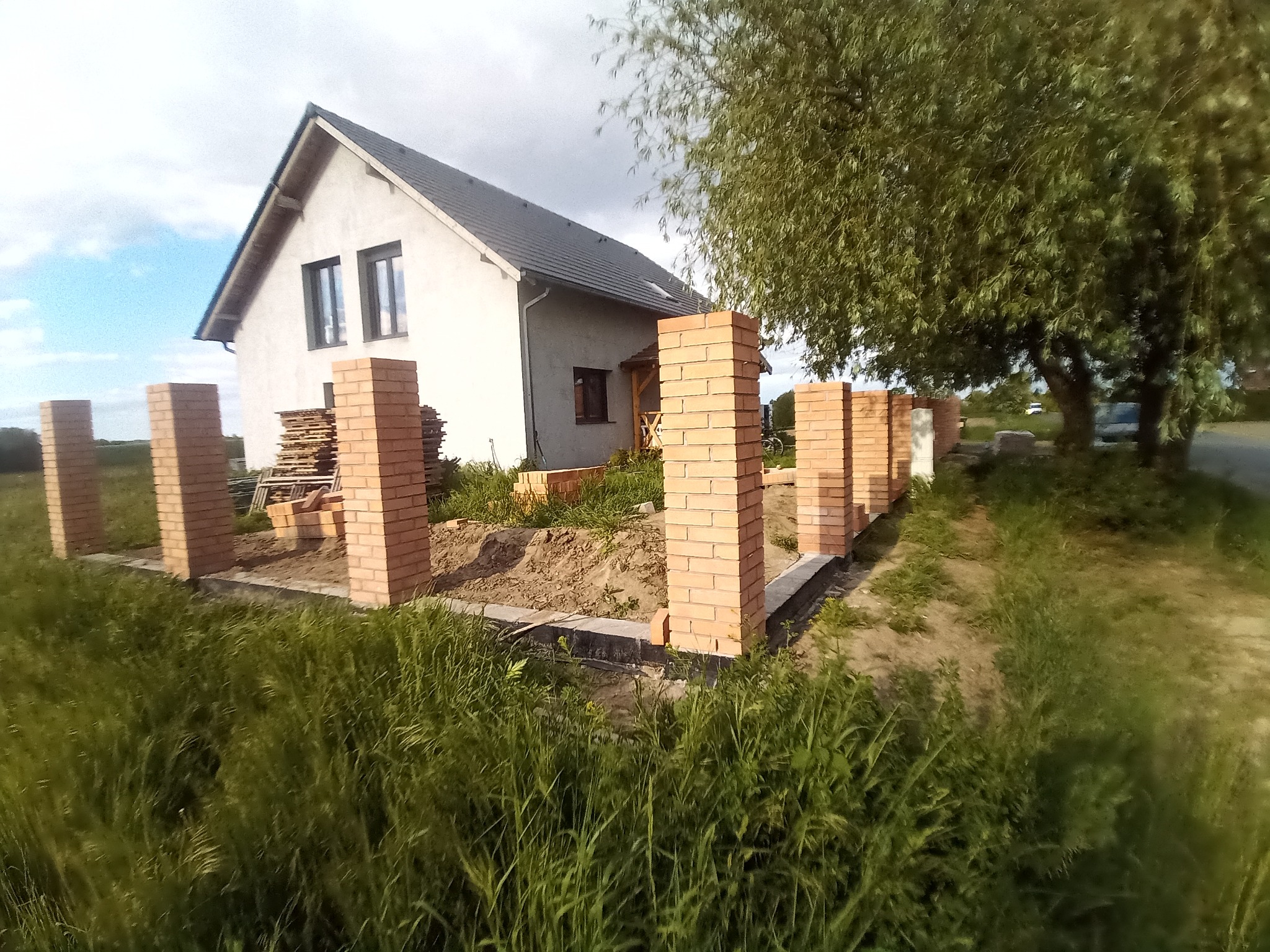 Budowa ogrodzenia z cegły klinkierowej przy domu jednorodzinnym. Widoczne słupki ogrodzeniowe w trakcie wznoszenia, zarys fundamentów i materiały budowlane.