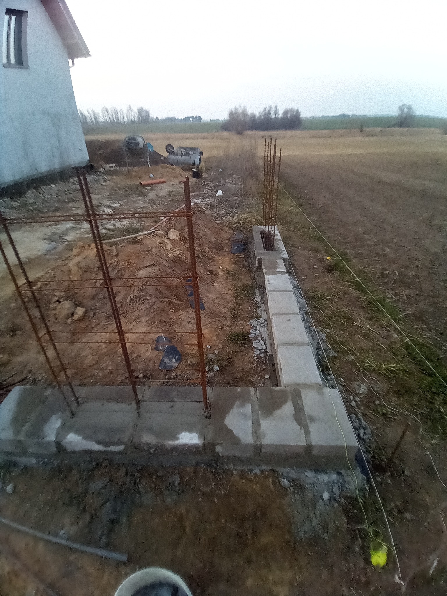 Wznoszenie fundamentów z betonu i zbrojenia na działce, widok z góry. Częściowo ułożone bloczki fundamentowe, w tle budynek i krajobraz polny.