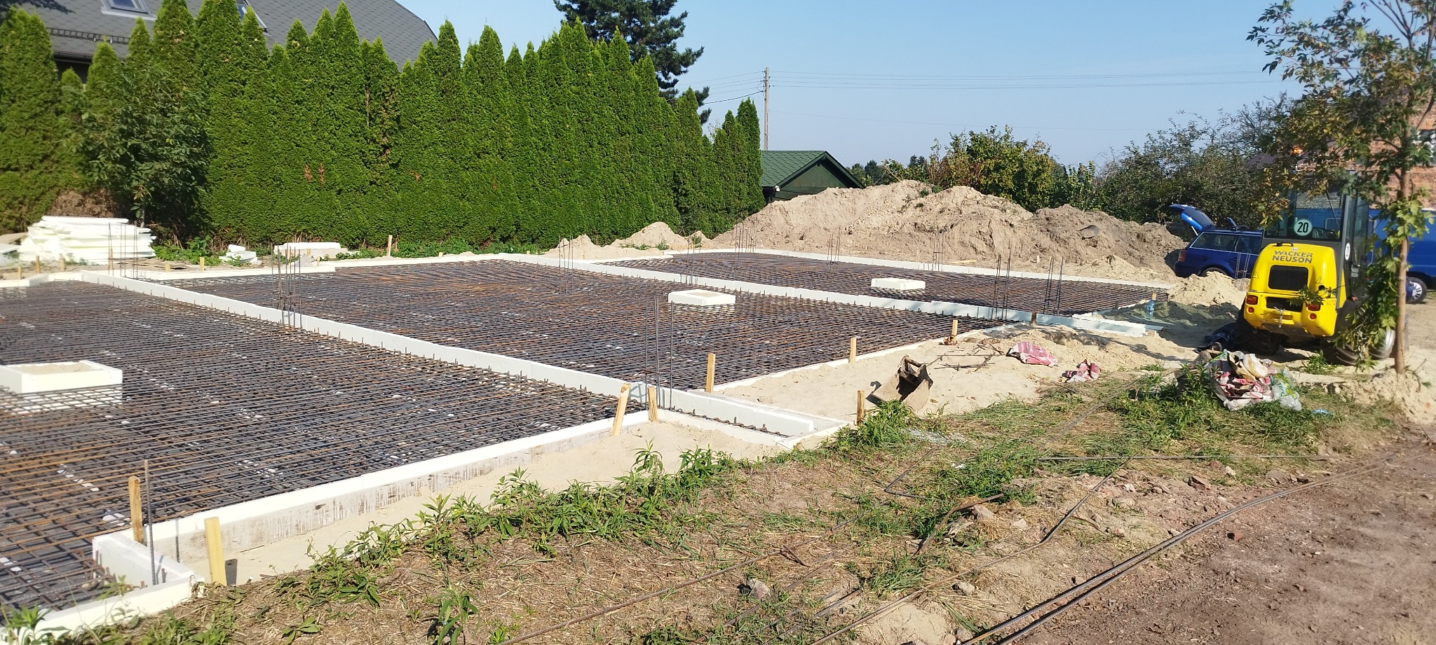 Zbrojenie fundamentów domu jednorodzinnego, widoczne stalowe pręty tworzące siatkę, białe szalunki, stos styropianu, na drugim planie koparka i samochody.