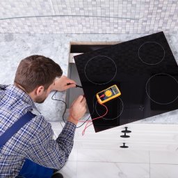 Pawel Ponomarjov - Elektryk w niebieskim kombinezonie sprawdza napięcie płyty indukcyjnej za pomocą multimetru; widok z góry na blat kuchenny z szarego marmuru i białe szafki.