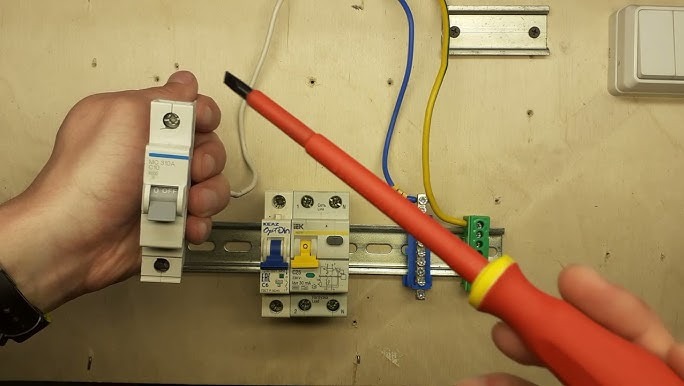 Instalacja elektryczna: ręka trzyma wyłącznik nad szyną DIN z zamontowanymi zabezpieczeniami, w tle widoczne przewody i włącznik światła na drewnianej płycie.