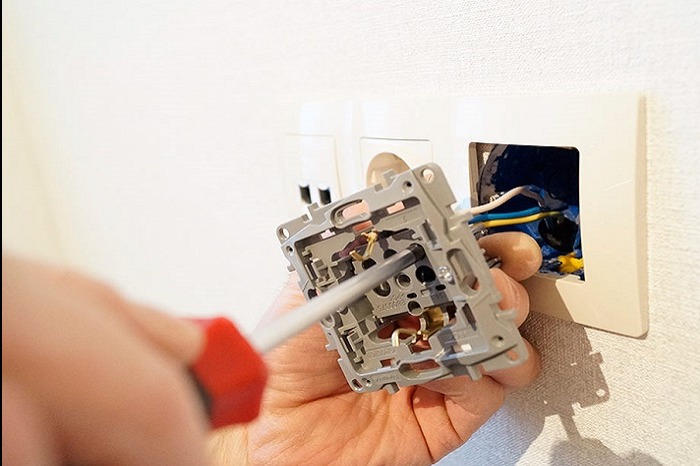 Elektryk przykręca gniazdko elektryczne do puszki w ścianie za pomocą śrubokręta z czerwoną rączką, widoczne przewody w kolorach niebieskim, żółtym i białym.