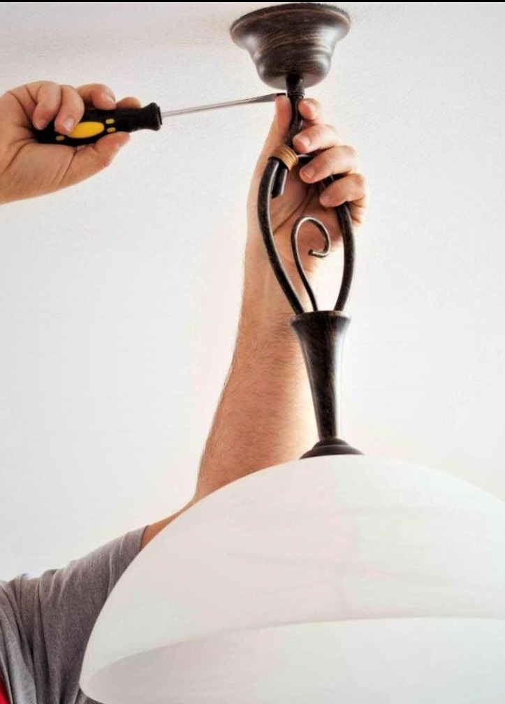 Instalacja ozdobnej lampy sufitowej z kloszem w stylu retro; elektryk dokręca śrubę mocującą oprawę do sufitu za pomocą śrubokręta z żółto-czarną rękojeścią.