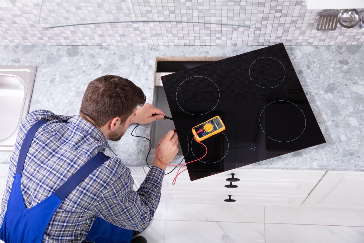 Elektryk w niebieskim kombinezonie sprawdza napięcie płyty indukcyjnej za pomocą multimetru; widok z góry na blat kuchenny z szarego marmuru i białe szafki.