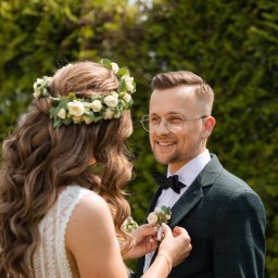 Okiem Alicji - Panna młoda przypina butonierkę do zielonego garnituru w kratę pana młodego, oboje uśmiechnięci na tle zieleni.