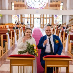 Okiem Alicji - Uśmiechnięta para młoda w kościele, panna młoda z bukietem i pierścionkiem, pan młody w niebieskim garniturze, dekoracje kwiatowe na ławkach, goście w tle.