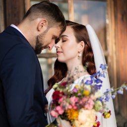 Okiem Alicji - Nowożeńcy dotykają się czołami, panna młoda w welonie trzyma bukiet wielokolorowych kwiatów na tle drewnianej ramy okiennej.