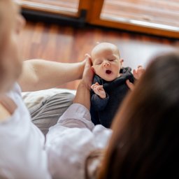 Okiem Alicji - Rodzice trzymają noworodka w czarnym ubranku, który ziewa z otwartymi ustami, na tle drewnianej podłogi i okna.