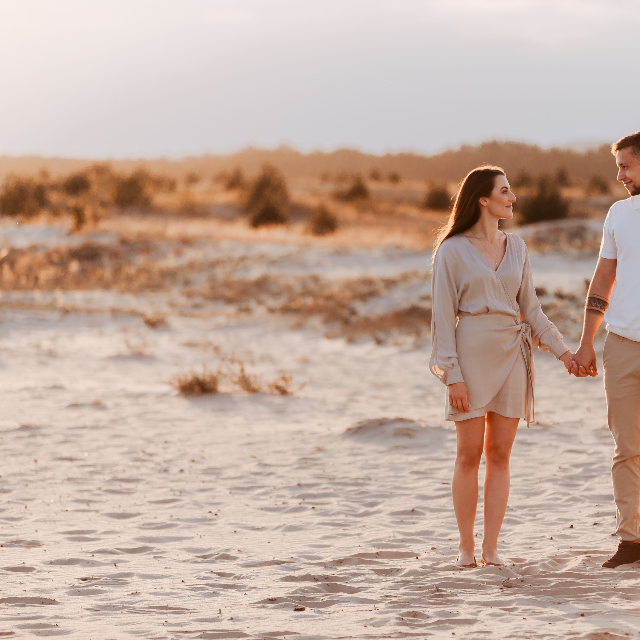 Para trzymająca się za ręce na piaszczystej plaży o zachodzie słońca, kobieta w beżowej sukience, mężczyzna z tatuażem na ramieniu.