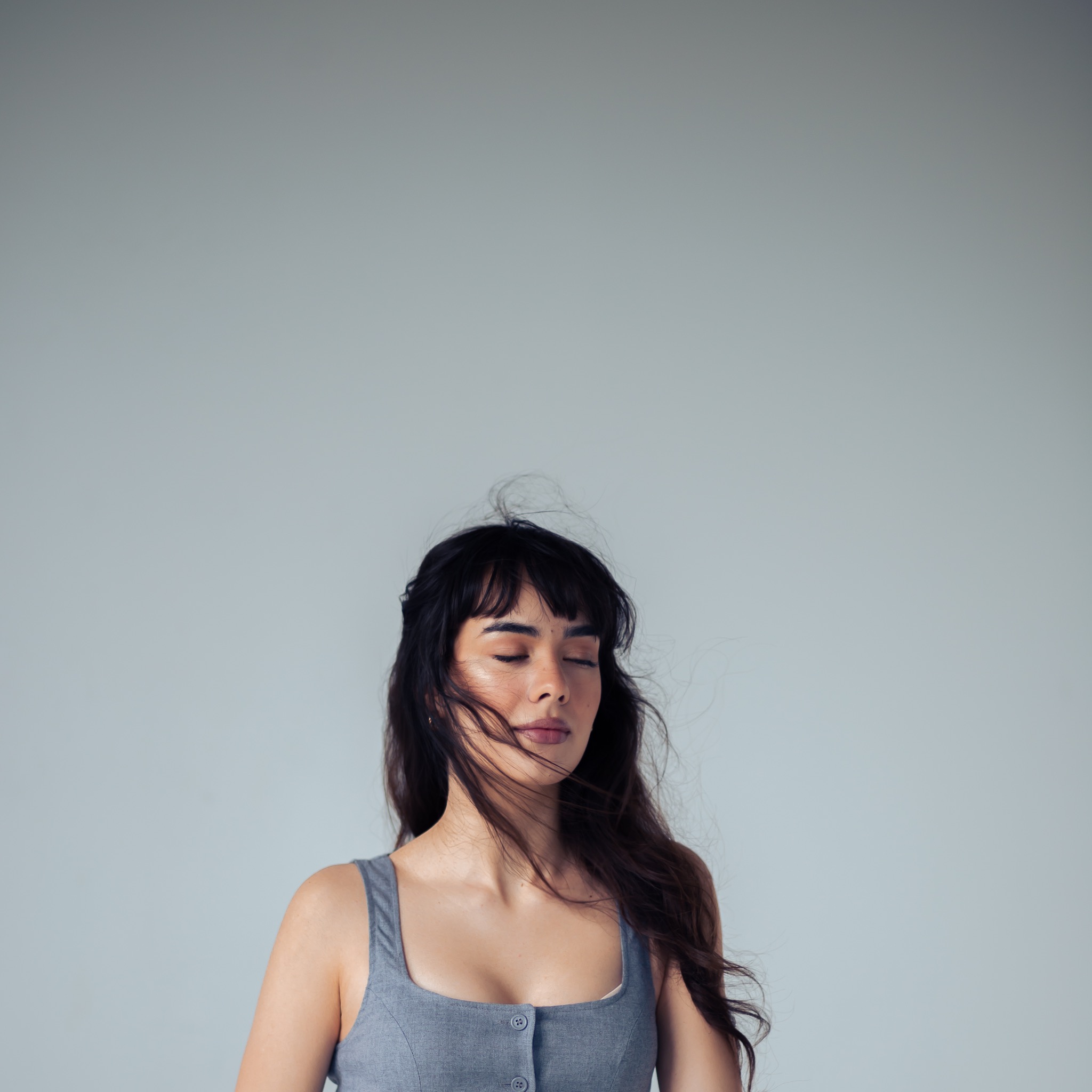 Portret kobiety z zamkniętymi oczami, włosy rozwiane, ubrana w szarą sukienkę na tle jasnoszarego studia.
