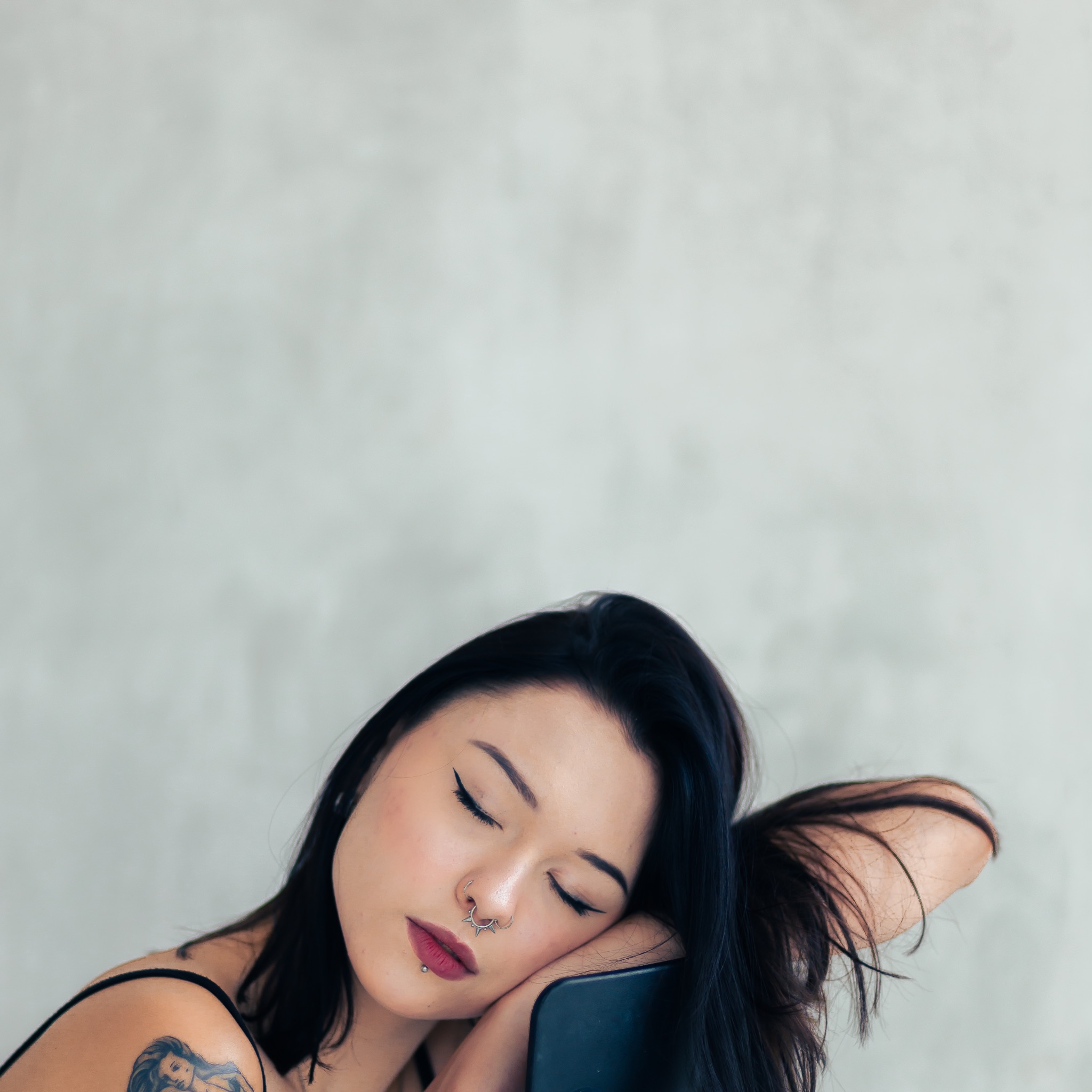 Portret kobiety z zamkniętymi oczami, opierającej głowę na krześle, widoczny tatuaż na ramieniu i liczne kolczyki na twarzy.