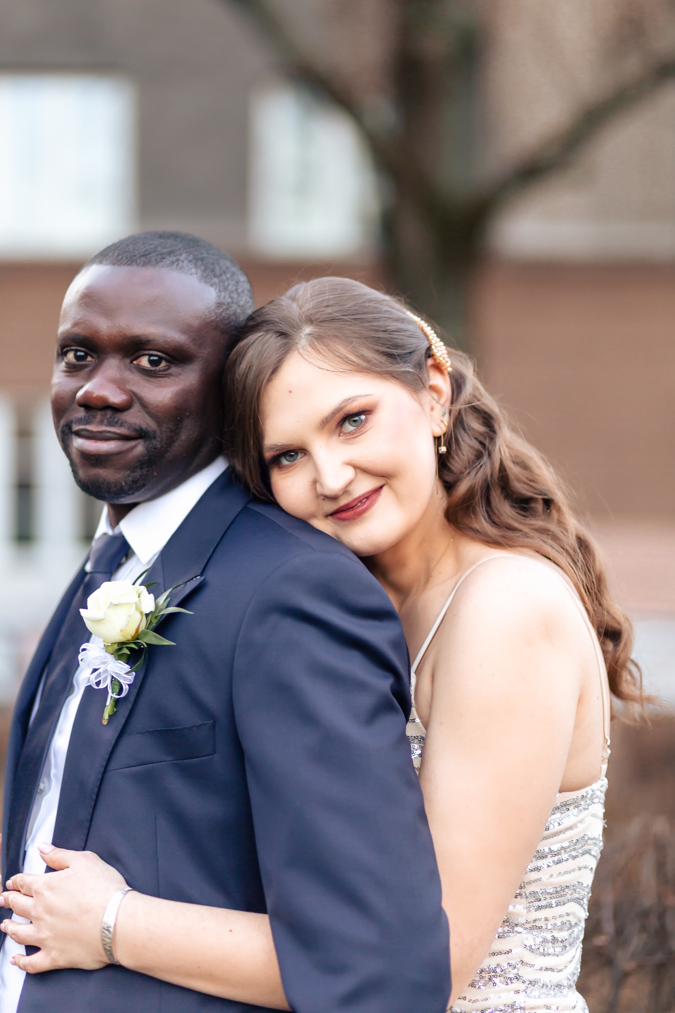 Uśmiechnięta para: mężczyzna w granatowym garniturze z butonierką z białej róży i kobieta w sukni z cekinami, obejmująca go czule.