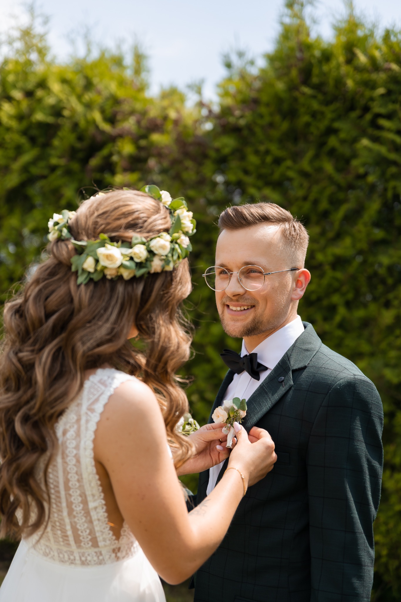 Panna młoda przypina butonierkę do zielonego garnituru w kratę pana młodego, oboje uśmiechnięci na tle zieleni.