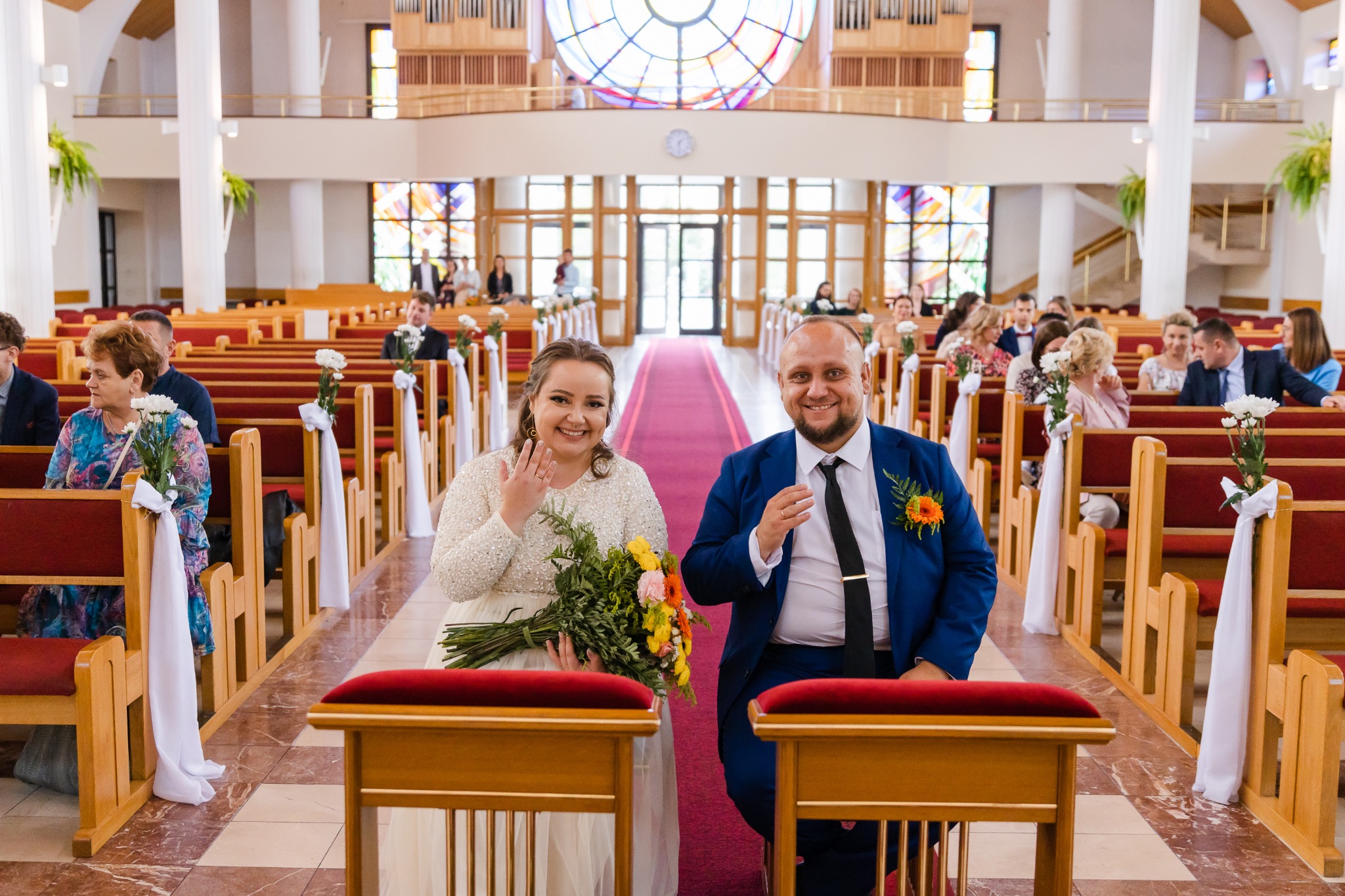 Uśmiechnięta para młoda w kościele, panna młoda z bukietem i pierścionkiem, pan młody w niebieskim garniturze, dekoracje kwiatowe na ławkach, goście w tle.