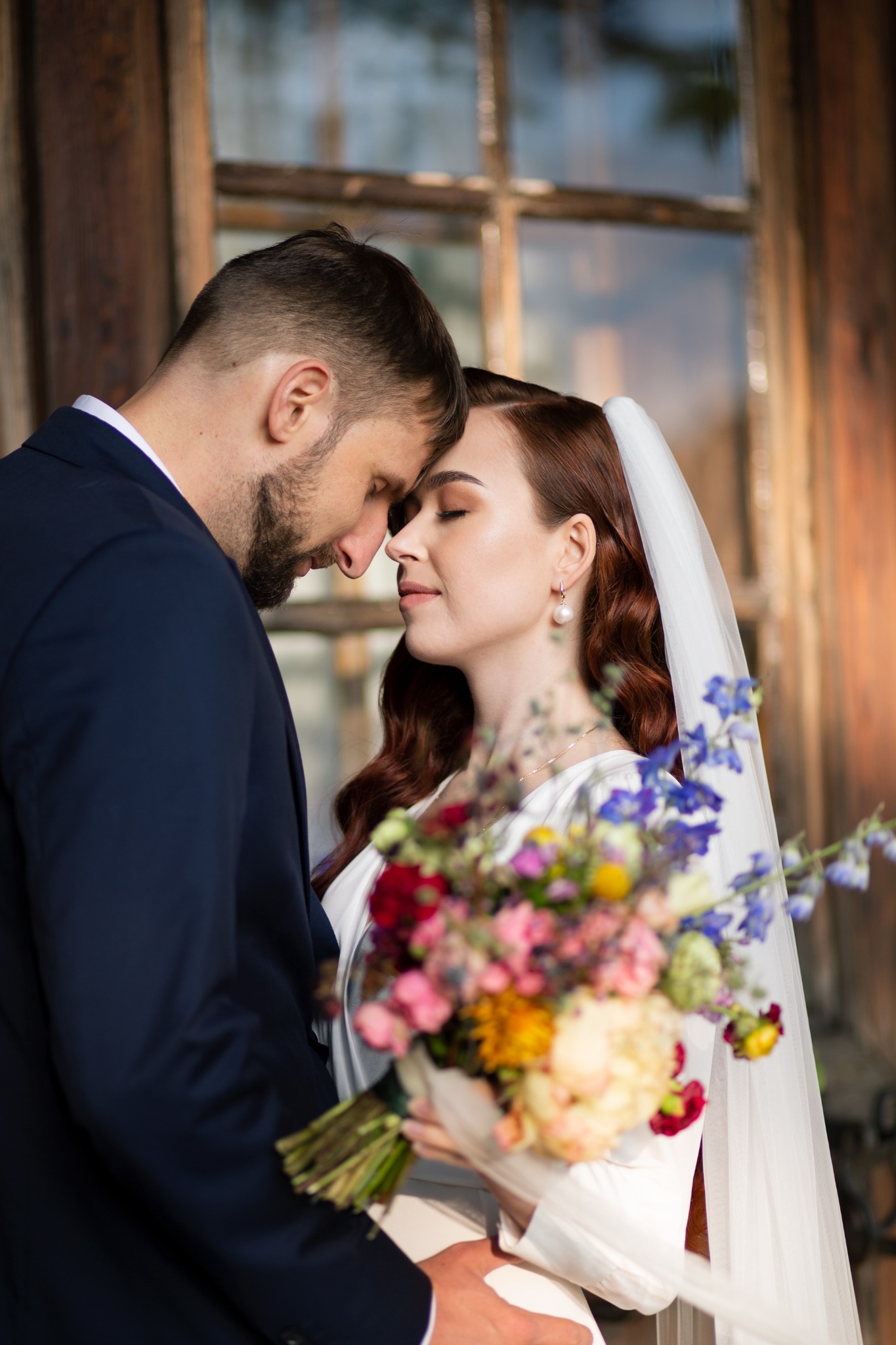 Nowożeńcy dotykają się czołami, panna młoda w welonie trzyma bukiet wielokolorowych kwiatów na tle drewnianej ramy okiennej.