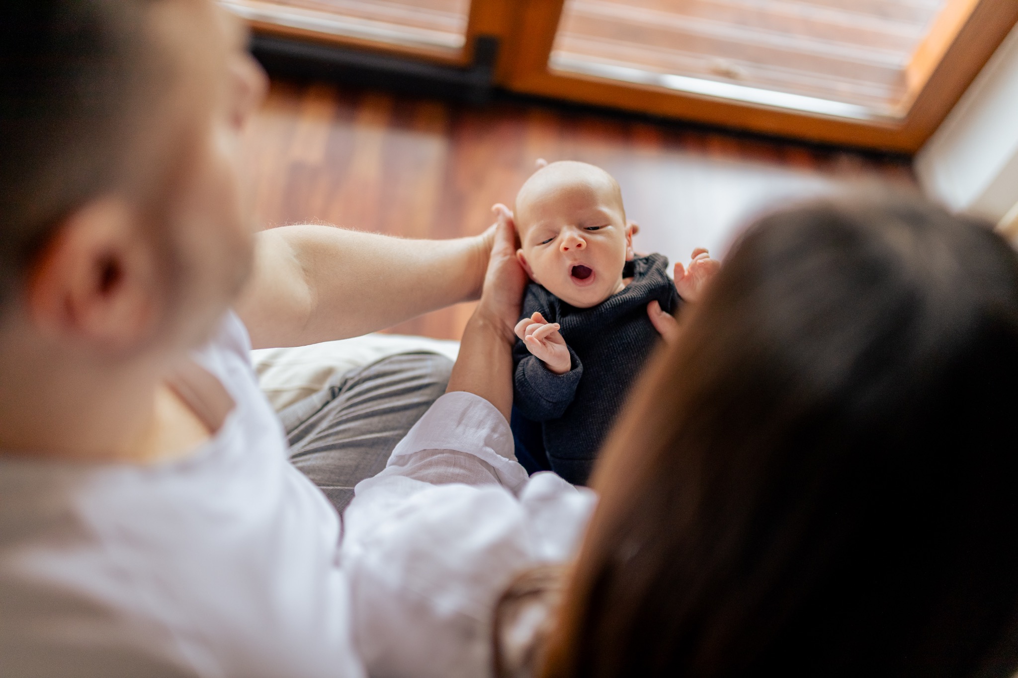 Rodzice trzymają noworodka w czarnym ubranku, który ziewa z otwartymi ustami, na tle drewnianej podłogi i okna.