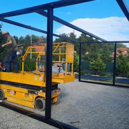 ZIBICREATELINE SPÓŁKA Z OGRANICZONĄ ODPOWIEDZIALNOŚCIĄ - Pracownik na podnośniku montuje metalową konstrukcję pergoli na żwirowym podłożu, widoczne narzędzia i elementy montażowe.