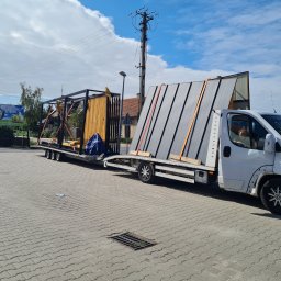 ZIBICREATELINE SPÓŁKA Z OGRANICZONĄ ODPOWIEDZIALNOŚCIĄ - Laweta z metalową konstrukcją pergoli i panelami ogrodzeniowymi zabezpieczonymi pasami transportowymi, zaparkowana na brukowanym placu.