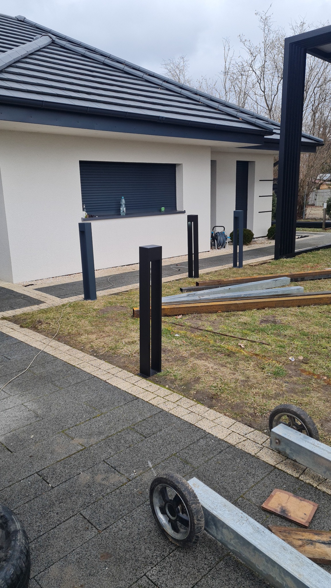Montaż nowoczesnego, minimalistycznego ogrodzenia z metalowych słupków w kolorze antracytowym, widoczne elementy konstrukcyjne i narzędzia na trawniku, w tle dom z szarą dachówką.