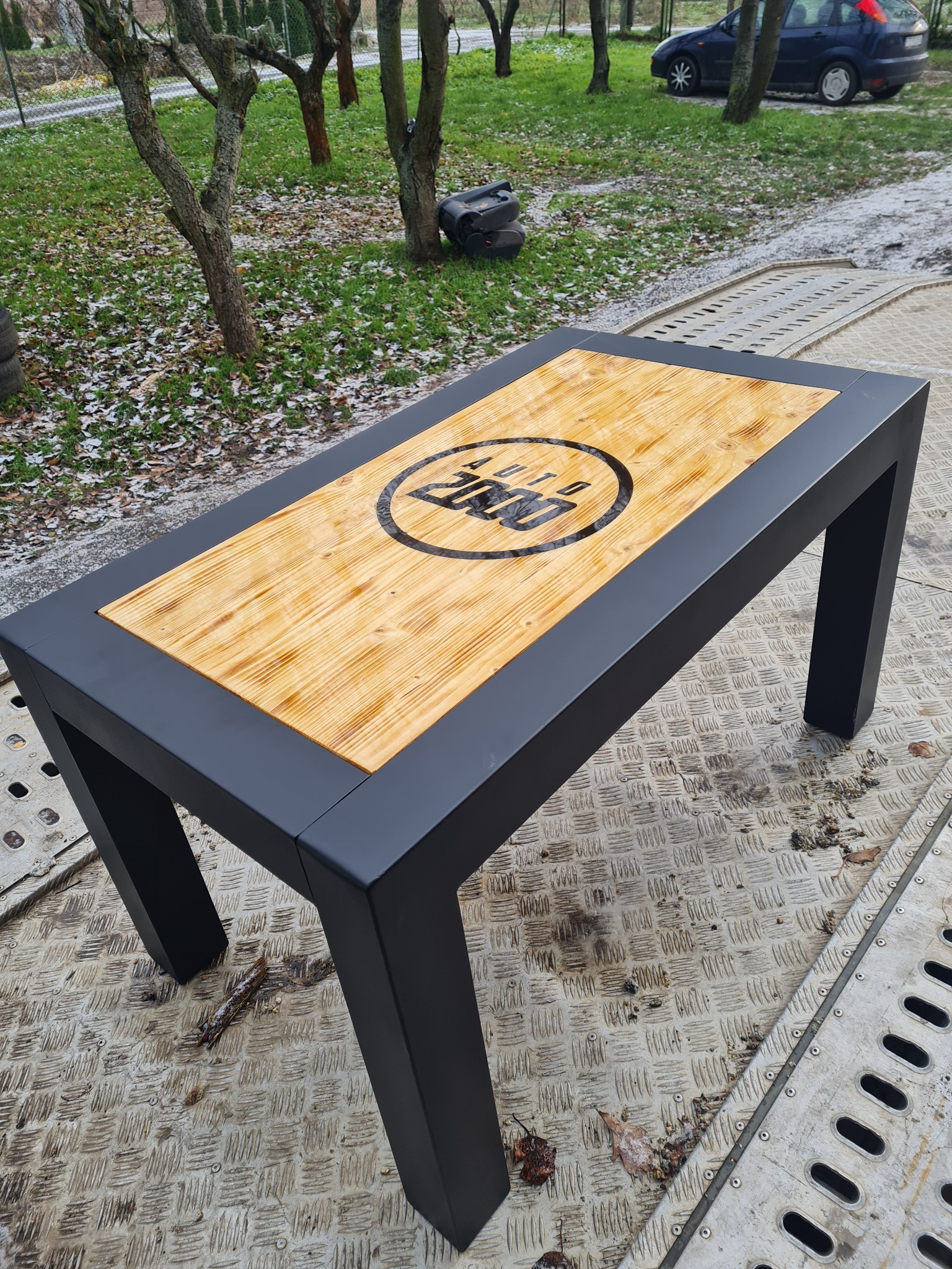 Metalowa pergola z drewnianym blatem z logo firmy, umieszczona na platformie transportowej w plenerze.