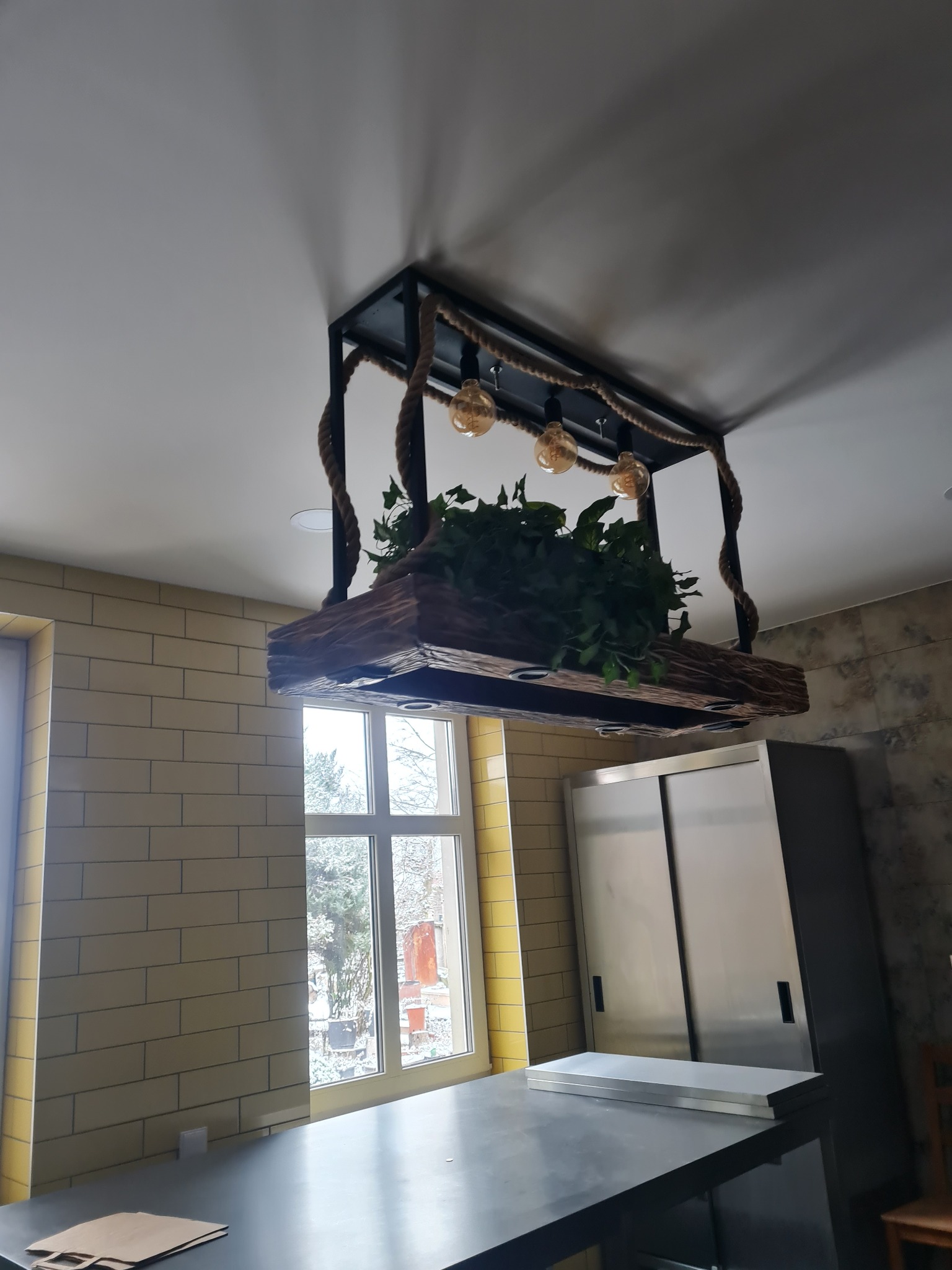 Dekoracyjna lampa sufitowa w stylu industrialnym: czarna metalowa rama z linami jutowymi, drewniana belka z roślinami i trzema żarówkami Edisona, widok z dołu, jasne wnętrze z oknem.