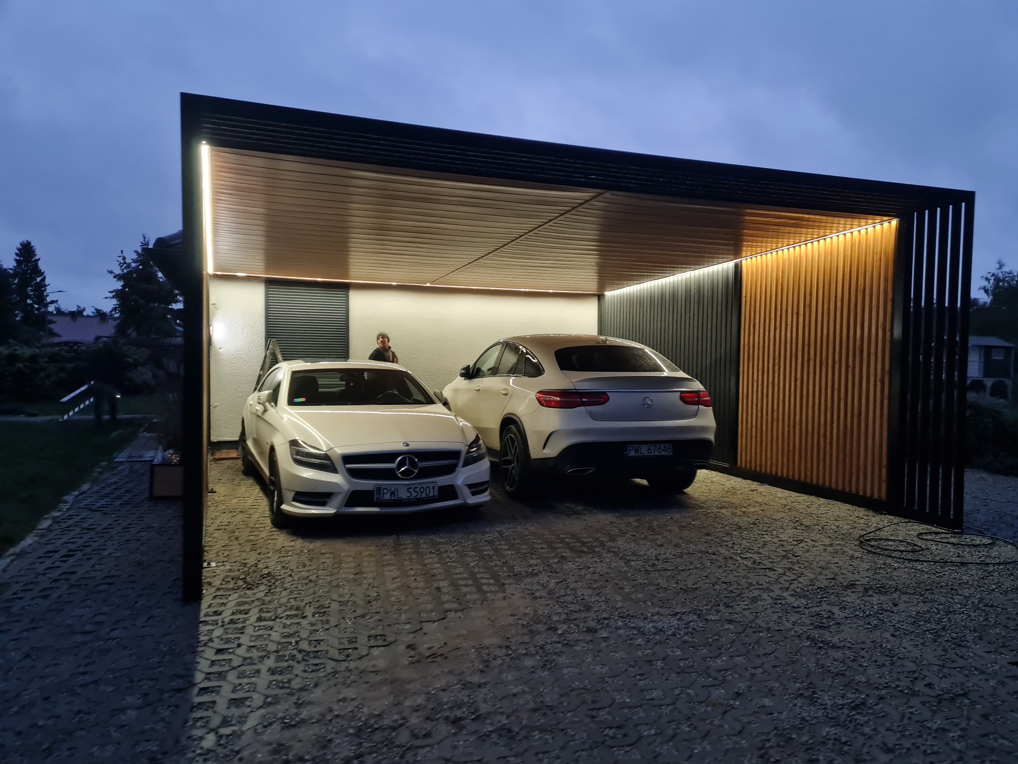 Nowoczesna wiata garażowa z oświetleniem LED, wykończona drewnianymi panelami, z dwoma białymi samochodami Mercedes w środku, widok z zewnątrz na podjazd z ażurowych płyt.