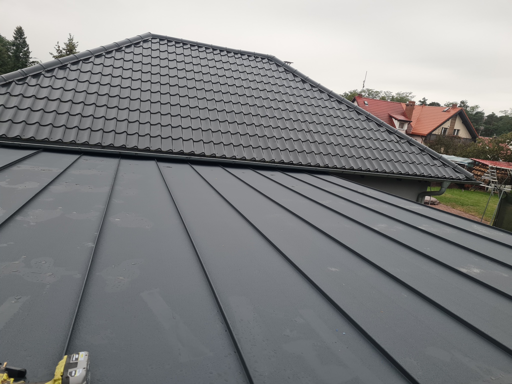 Fragment dachu z blachy na rąbek stojący i dachówki ceramicznej w kolorze grafitowym, widoczne narzędzie montażowe w lewym dolnym rogu, w tle dom z czerwoną dachówką.