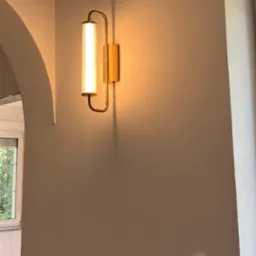 Ścienny kinkiet w stylu art deco z podłużnym, prążkowanym kloszem i złotą oprawą, zamontowany na gładkiej, beżowej ścianie z widocznym, okrągłym gniazdkiem elektrycznym.