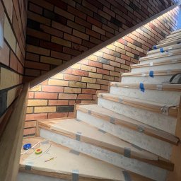 Diego Service - Montaż oświetlenia schodowego z ceglaną ścianą, widoczne przewody i narzędzia elektryczne na dolnym stopniu, zabezpieczone stopnie schodów.