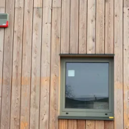 Zewnętrzny panel alarmowy z czerwoną obwódką zamontowany na drewnianej elewacji budynku obok okna z szarą ramą.