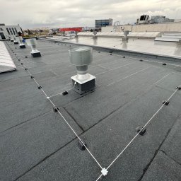 Diego Service - Płaski dach pokryty papą z zainstalowaną instalacją odgromową, widoczne metalowe kominki wentylacyjne i przewody odgromowe z uchwytami, w tle budynki przemysłowe i pochmurne niebo.