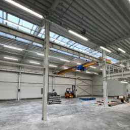 Wnętrze hali magazynowej z suwnicą, oświetleniem LED i wózkiem widłowym, widoczne okna dachowe.