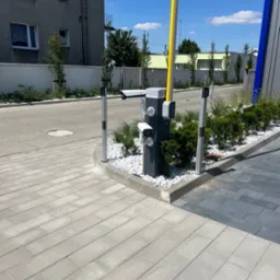 Automatyczna szlaban z żółtą belką w pozycji pionowej, kamery monitoringu na słupku, otoczenie wyłożone kostką brukową i ozdobione białymi kamieniami oraz zielenią.