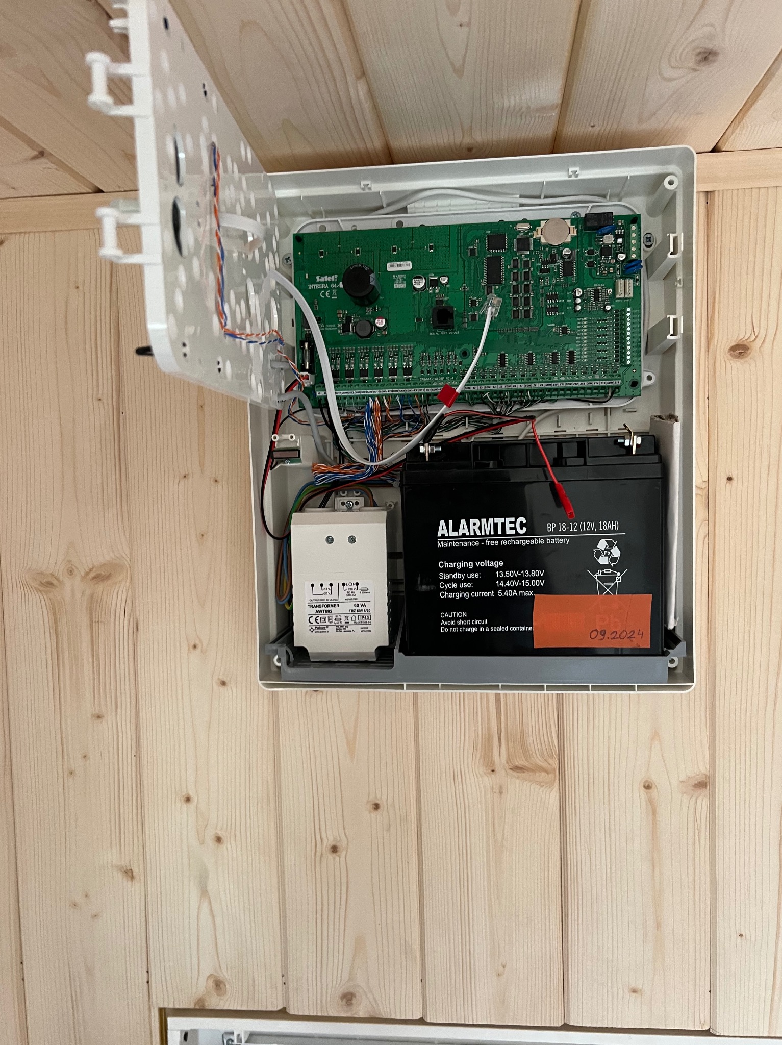 Otwarta obudowa centrali alarmowej z widoczną płytą główną, akumulatorem ALARMTEC i transformatorem, zamontowana na drewnianej ścianie z boazerii.