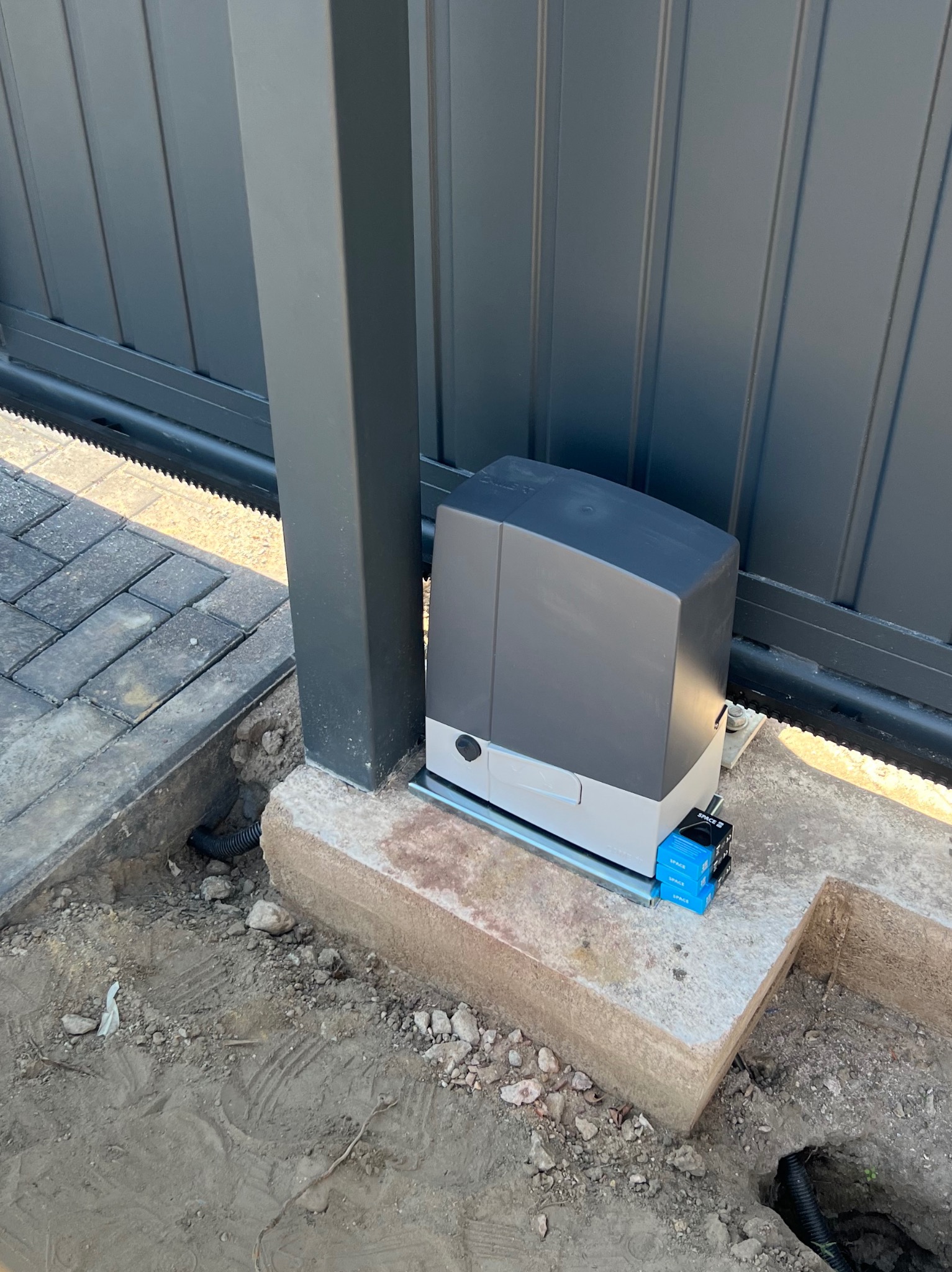 Szary automat do bramy przesuwnej zamontowany na betonowej podstawie, widoczna listwa zębata i skrzynka sterująca, obok kostka brukowa i ziemia.