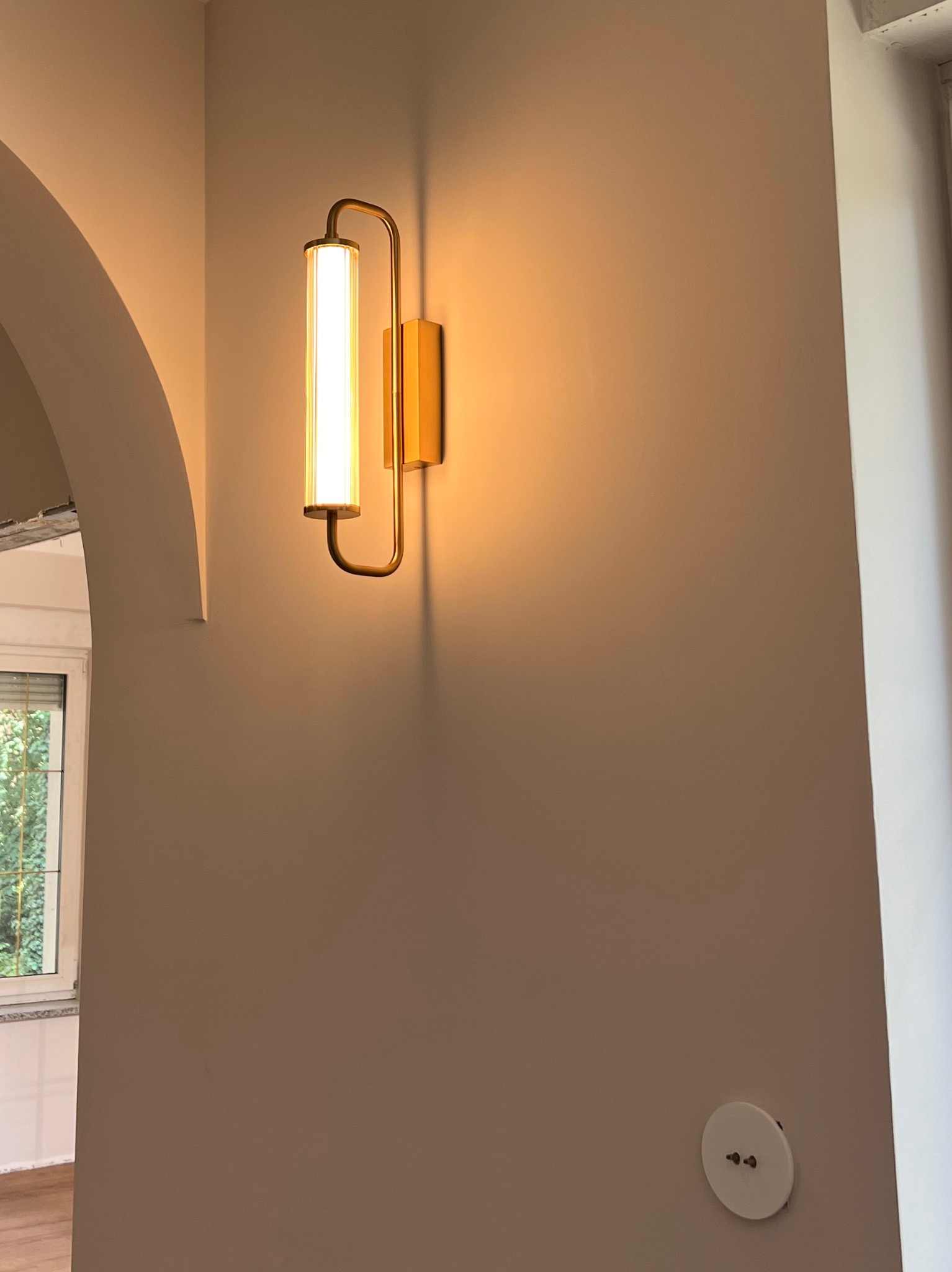 Ścienny kinkiet w stylu art deco z podłużnym, prążkowanym kloszem i złotą oprawą, zamontowany na gładkiej, beżowej ścianie z widocznym, okrągłym gniazdkiem elektrycznym.