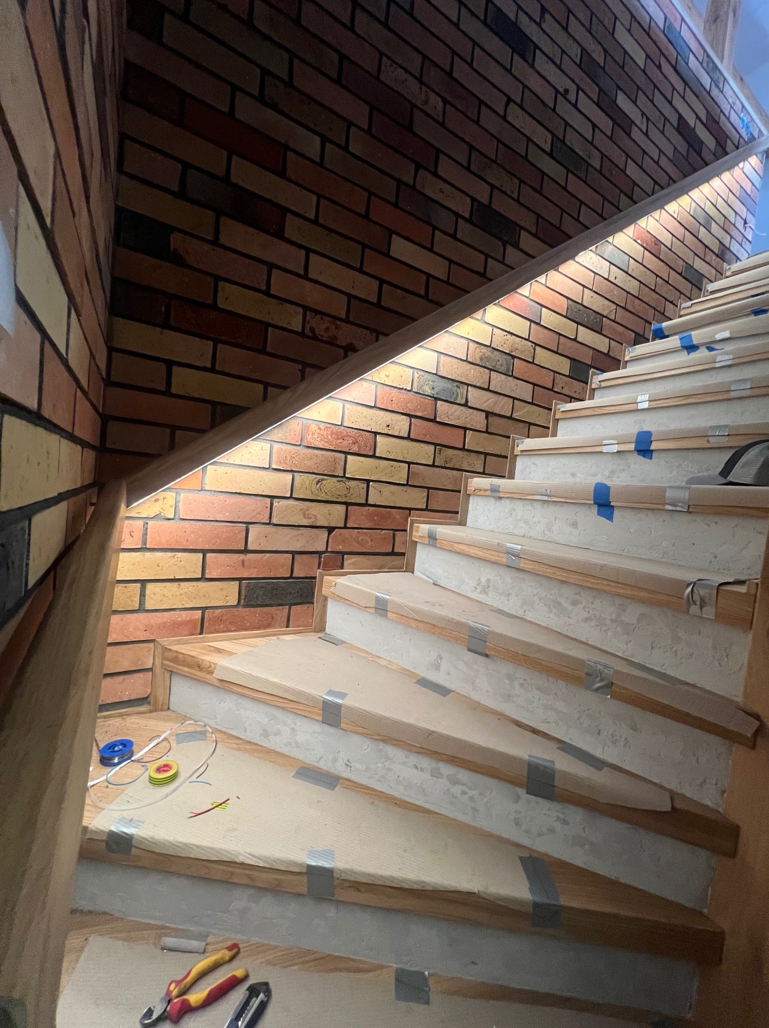 Montaż oświetlenia schodowego z ceglaną ścianą, widoczne przewody i narzędzia elektryczne na dolnym stopniu, zabezpieczone stopnie schodów.