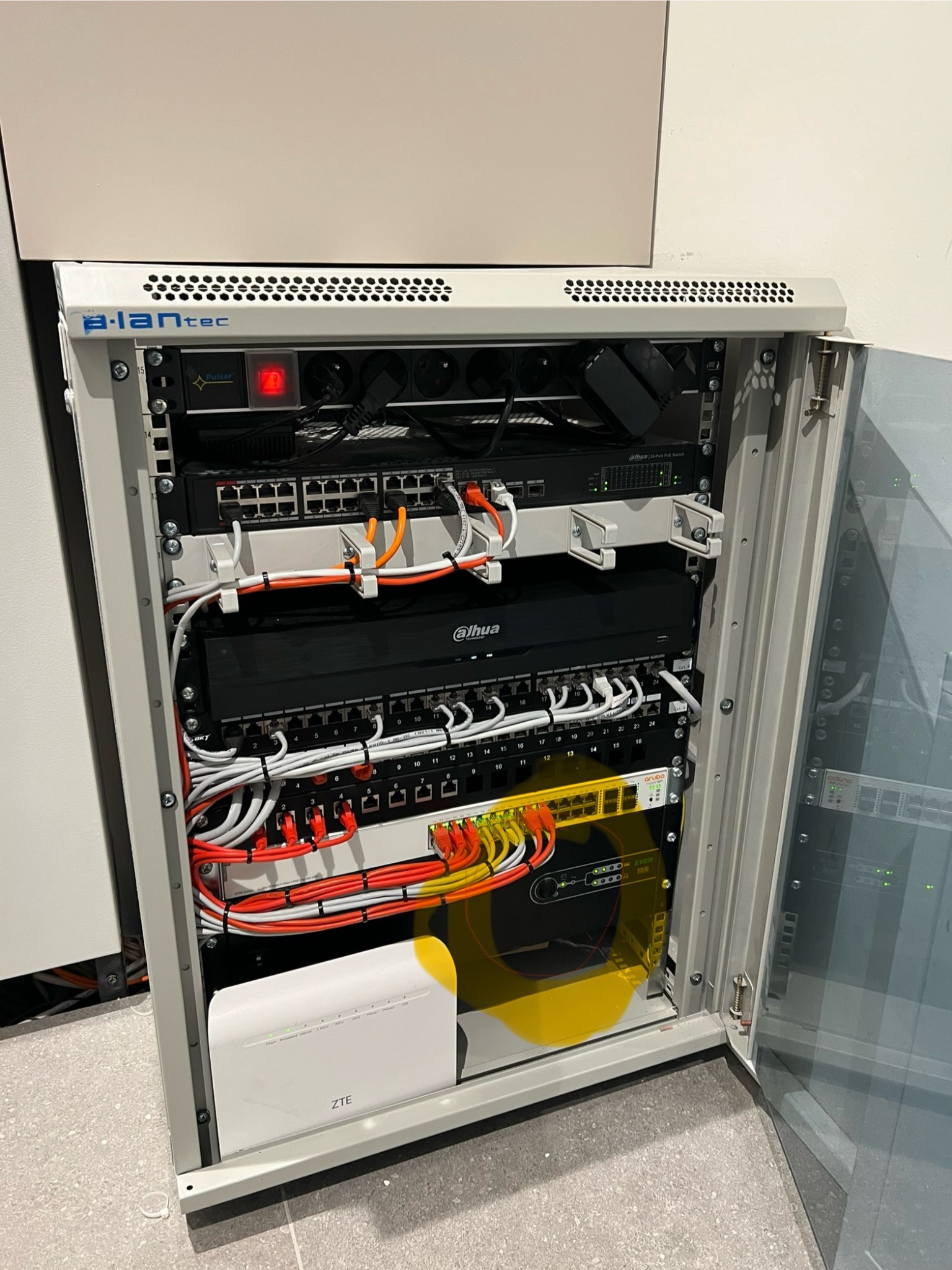 Otwarta szafa rack z zainstalowanym sprzętem sieciowym: switche, router ZTE i patch panel Dahua, okablowanie uporządkowane kolorowymi przewodami. Widoczne logo a.lantec na obudowie szafy.