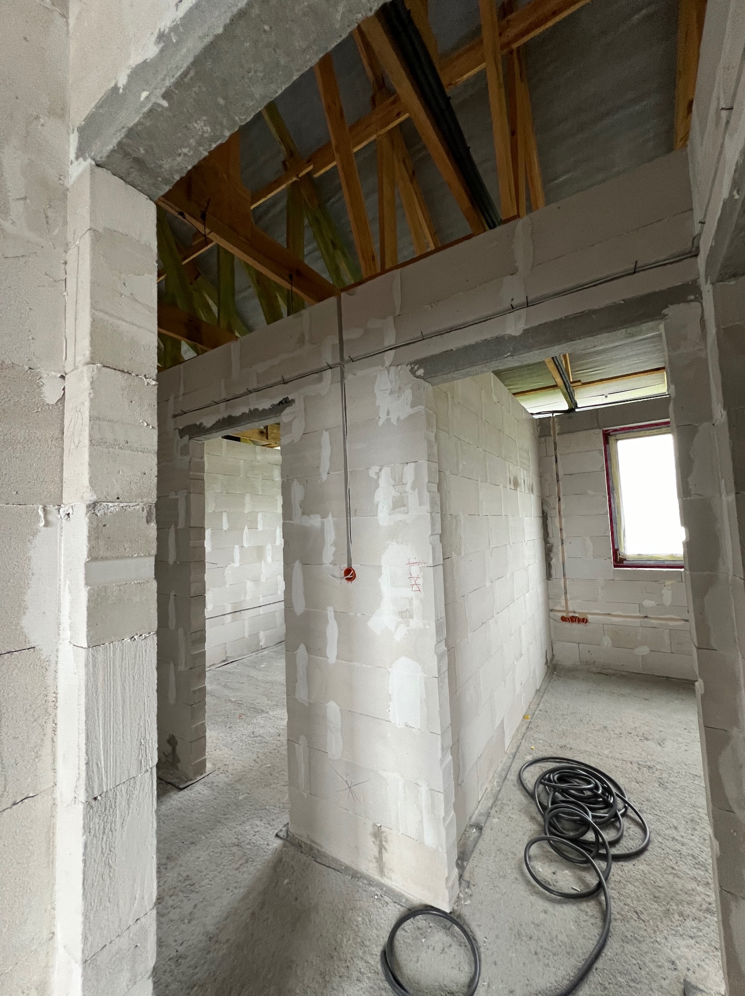 Wnętrze surowego domu w trakcie budowy z widocznymi przewodami elektrycznymi, ścianami z bloczków i drewnianą konstrukcją dachu.